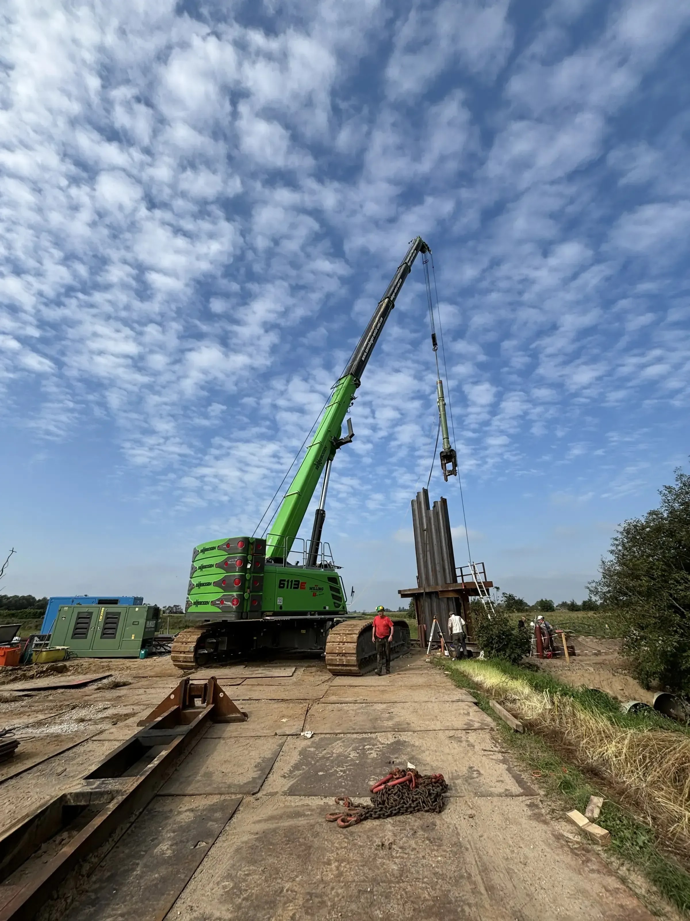 Grüner Raupenkran bringt Spundwände auf Baustelle ein, Arbeiter mit Helm daneben